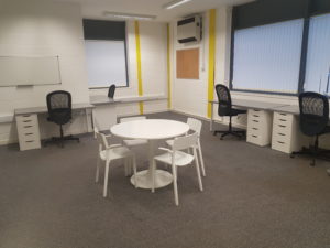 Inside view of the Minehead office space. There are desks facing two of the walls and a white round table with four chairs in the middle of the room. The windows are large and there are blinds.