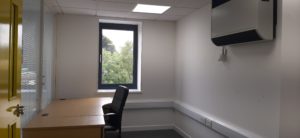 Inside view of a small office with a single desk and chair. There is a large window and an aircon unit on the wall.