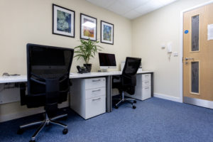 An office space with two desks side by side, blue carpet and art on the walls