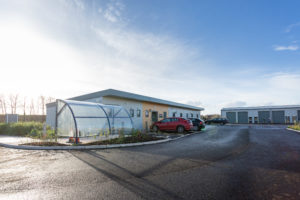 Wells building outside view. It's a one storey building and there are allocated parking spaces directly outside. There is also a covered bike storage area. A wide road leads to the next units.