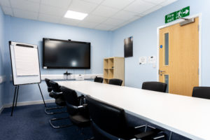 Highbridge meeting room. There is a long thin desk with 8 chairs around it. There is a screen mounted on the end wall, with a floor standing flip chart whiteboard next to it.