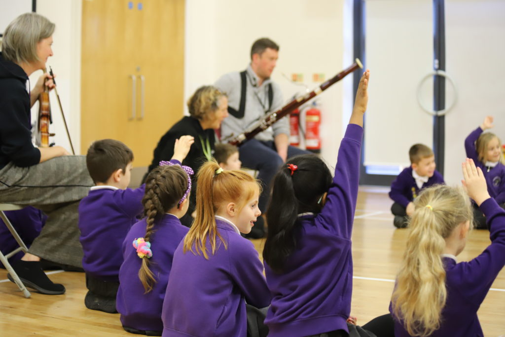 School children sat on the floor in a hall with musicians holding instruments.