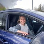 Lucy is sitting in a blue car with the window down, smiling at the camera