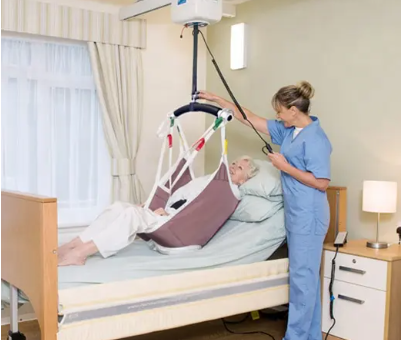 Carer helps elderly woman into bed using a hoist.