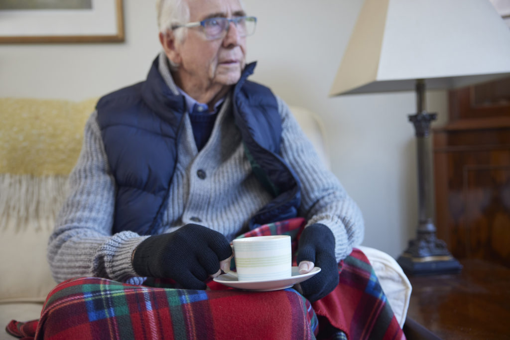 Senior man wearing extra clothes with hot drink trying to keep warm at home in energy crisis.