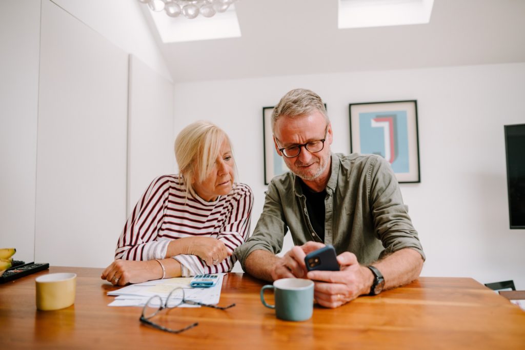 Man and woman are sat at a table looking at a phone. There is paperwork, a calculator and a pair of glasses on the table. They are discussing finances.