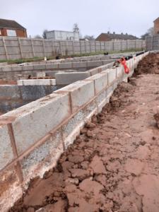 Construction site with partially built brick wall on muddy ground. Background includes a wooden fence, residential houses, and a red pipe.