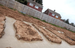 A construction site with trenches in the ground, filled with water. Two houses and a wooden fence are visible in the background.